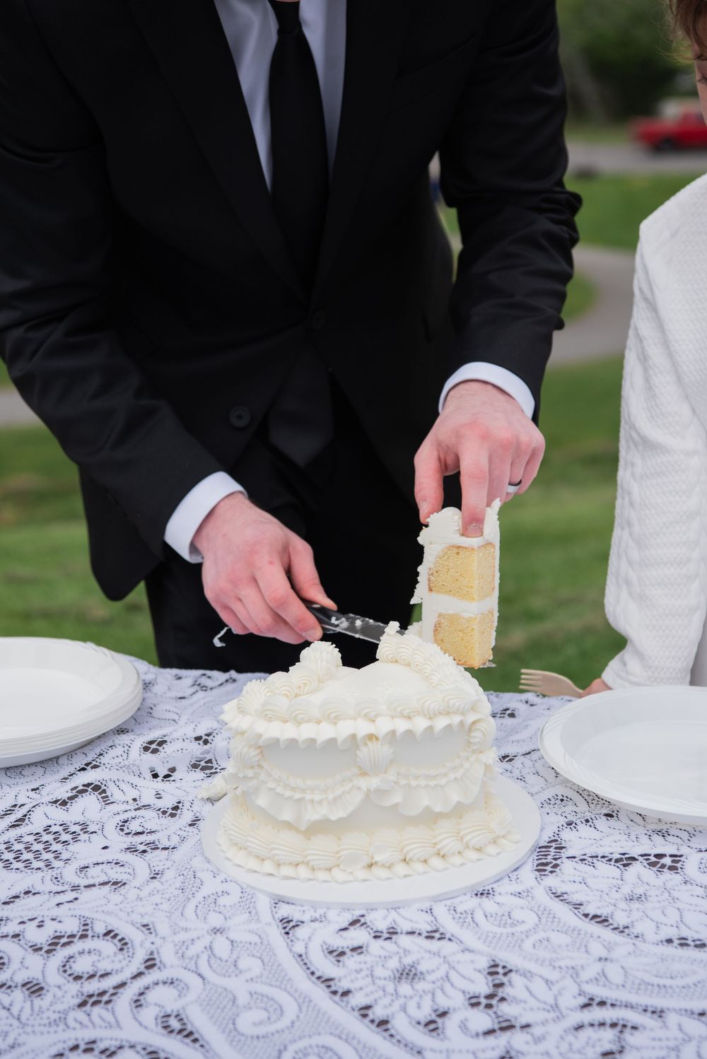 Couple cutting their white tiered wedding cake together on an outdoor table with lace tablecloth.