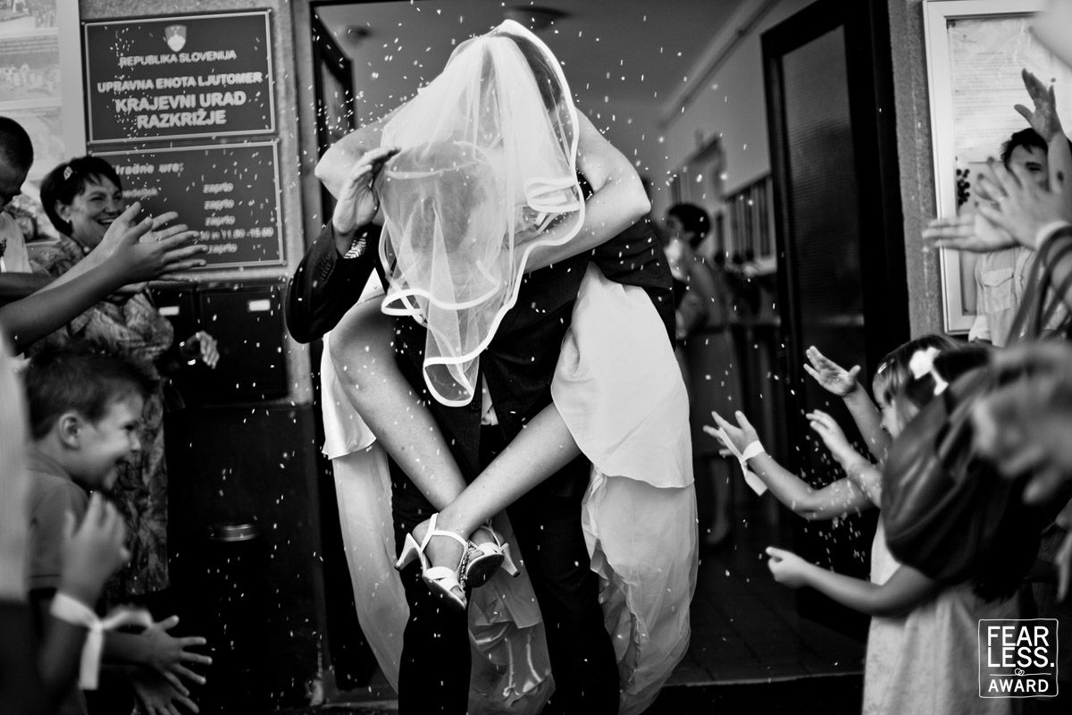A newlywed couple shares a celebratory kiss while guests cheer at a black and white wedding reception moment.