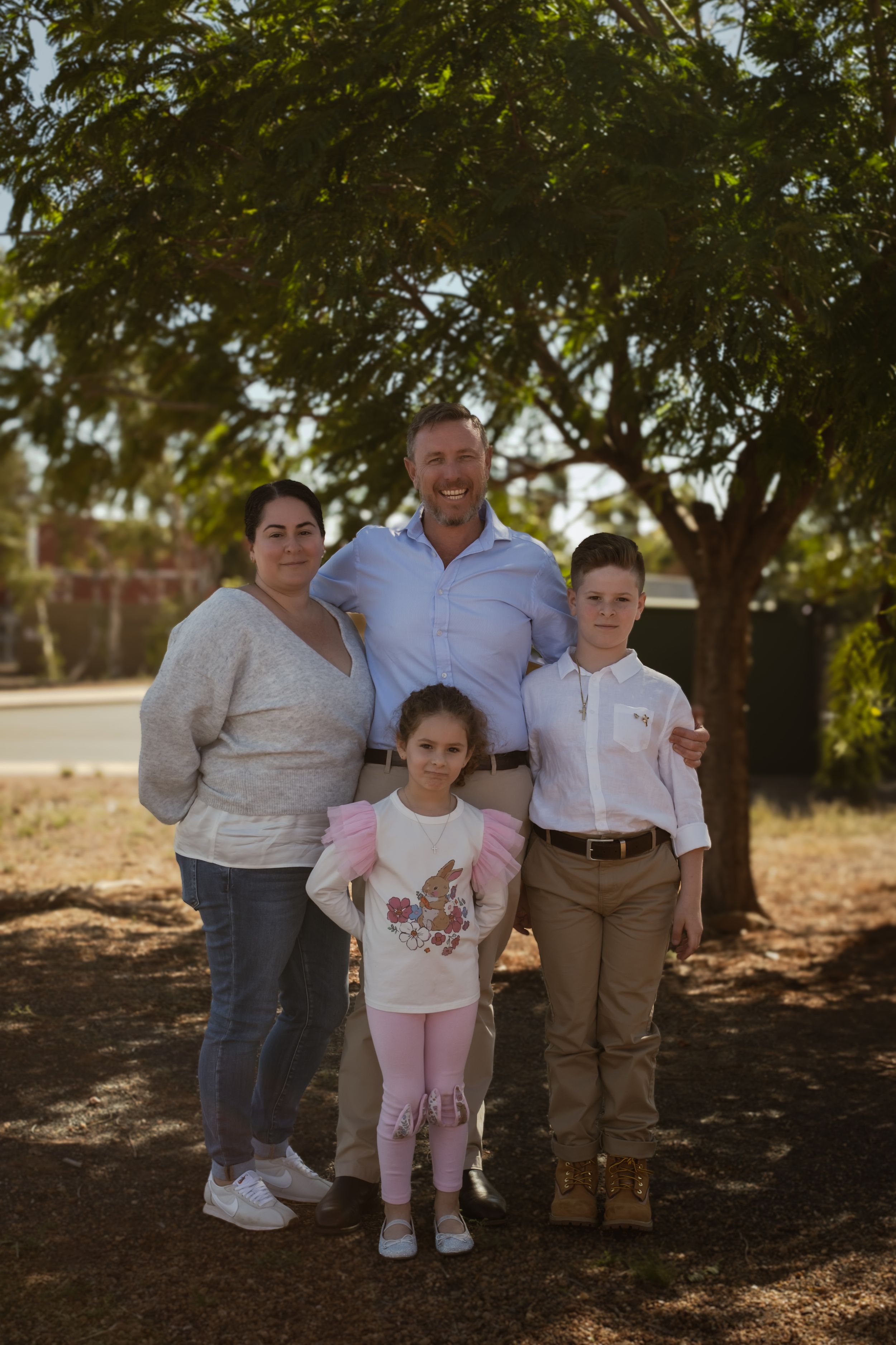 A casual family portrait taken under the shade of large trees in an outdoor setting.