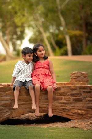 Two children sit barefoot on a brick wall in a garden setting, wearing a striped shirt and red polka dot dress.
