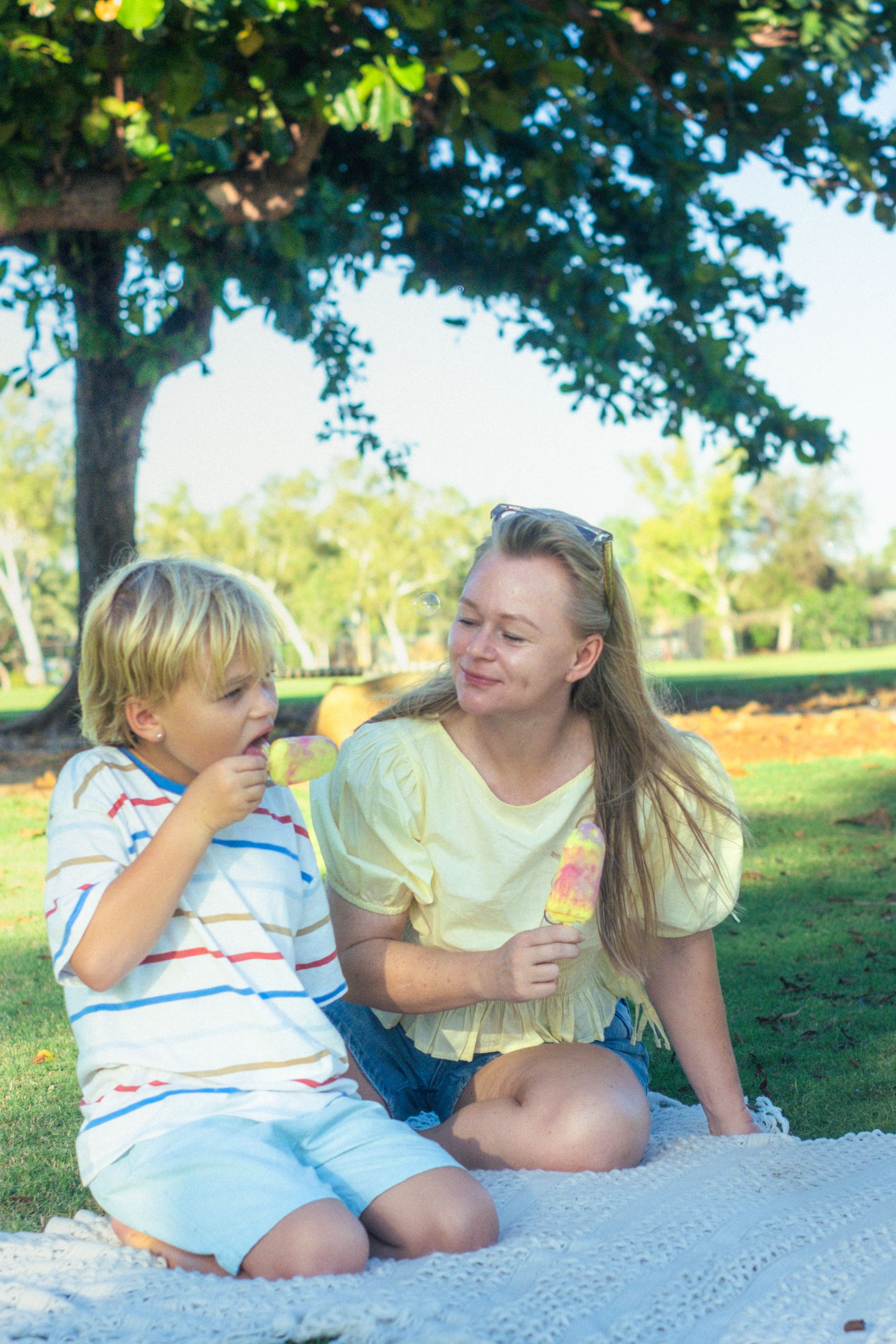 A mother and young boy enjoy ice cream together on a blanket in a sunny park under a large tree.