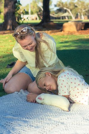 Two blonde girls bonding with a white guinea pig on a lace blanket in a sunny green park.