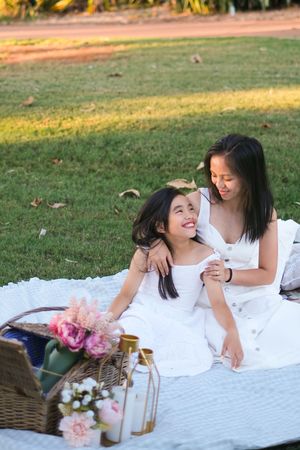 Friends relaxing on a white picnic blanket with pink flowers and decorative lanterns in a park setting.