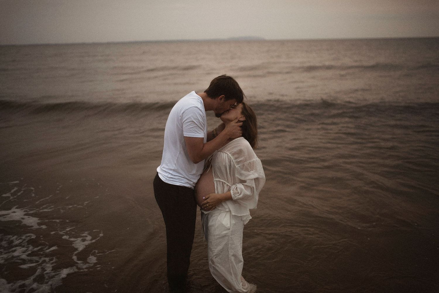 Two people embrace in a romantic moment on a moody beach at dusk, creating a dreamy atmospheric scene.