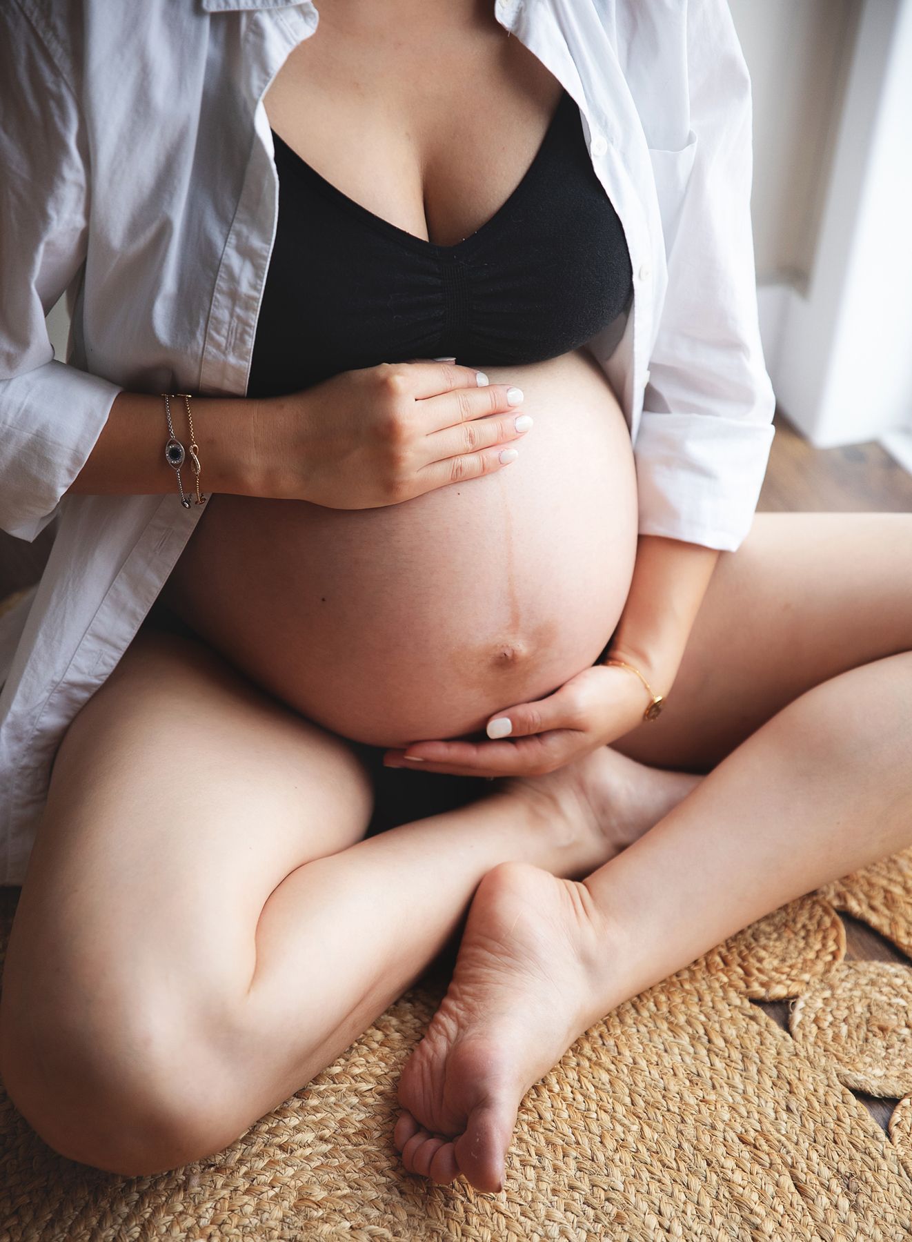 Close-up shot of a pregnant belly wearing a black bra and white shirt, sitting on a mat in natural light. At Maternity session in Sidcup