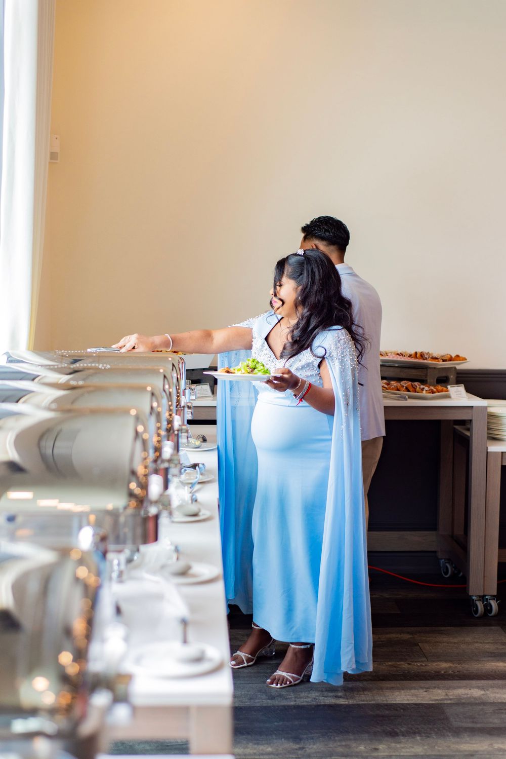 Expectant mother in flowing blue dress serves herself at an elegant catering buffet setup.