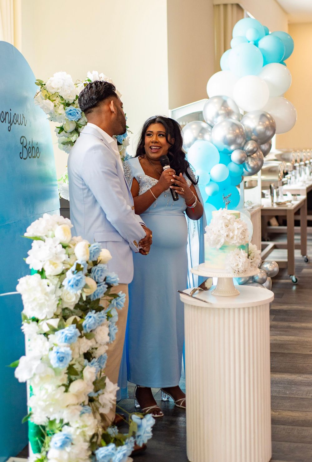 Baby shower celebration with blue and white balloon arch, floral decorations, and couple sharing joyful moment beside pillar display.