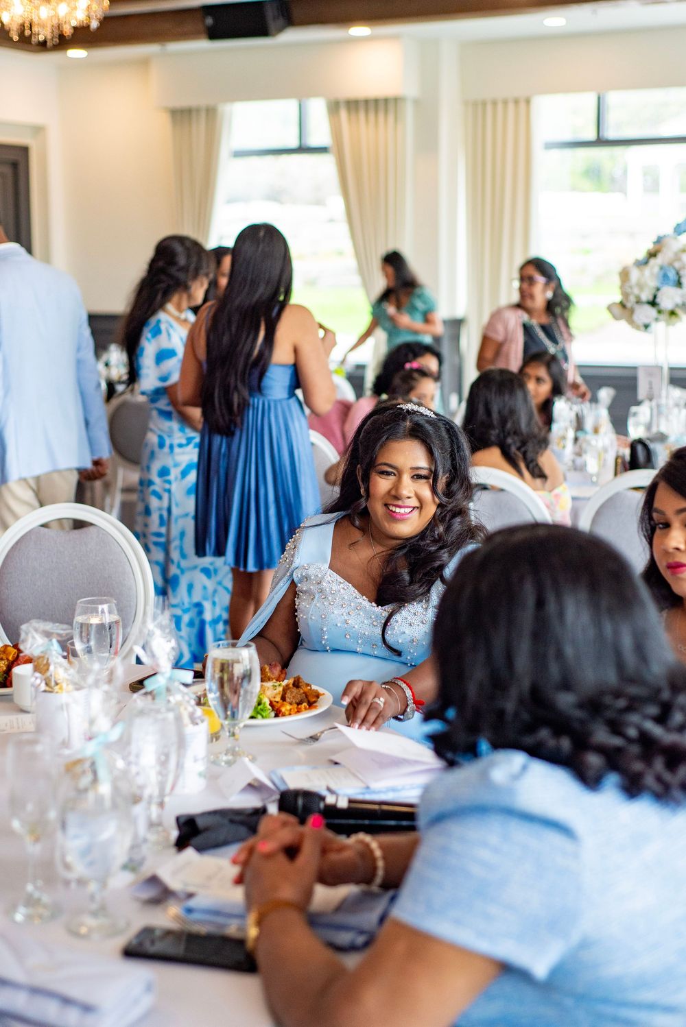 Guests in blue dresses enjoy a festive celebration at an elegant indoor reception with white tablecloths and large windows.