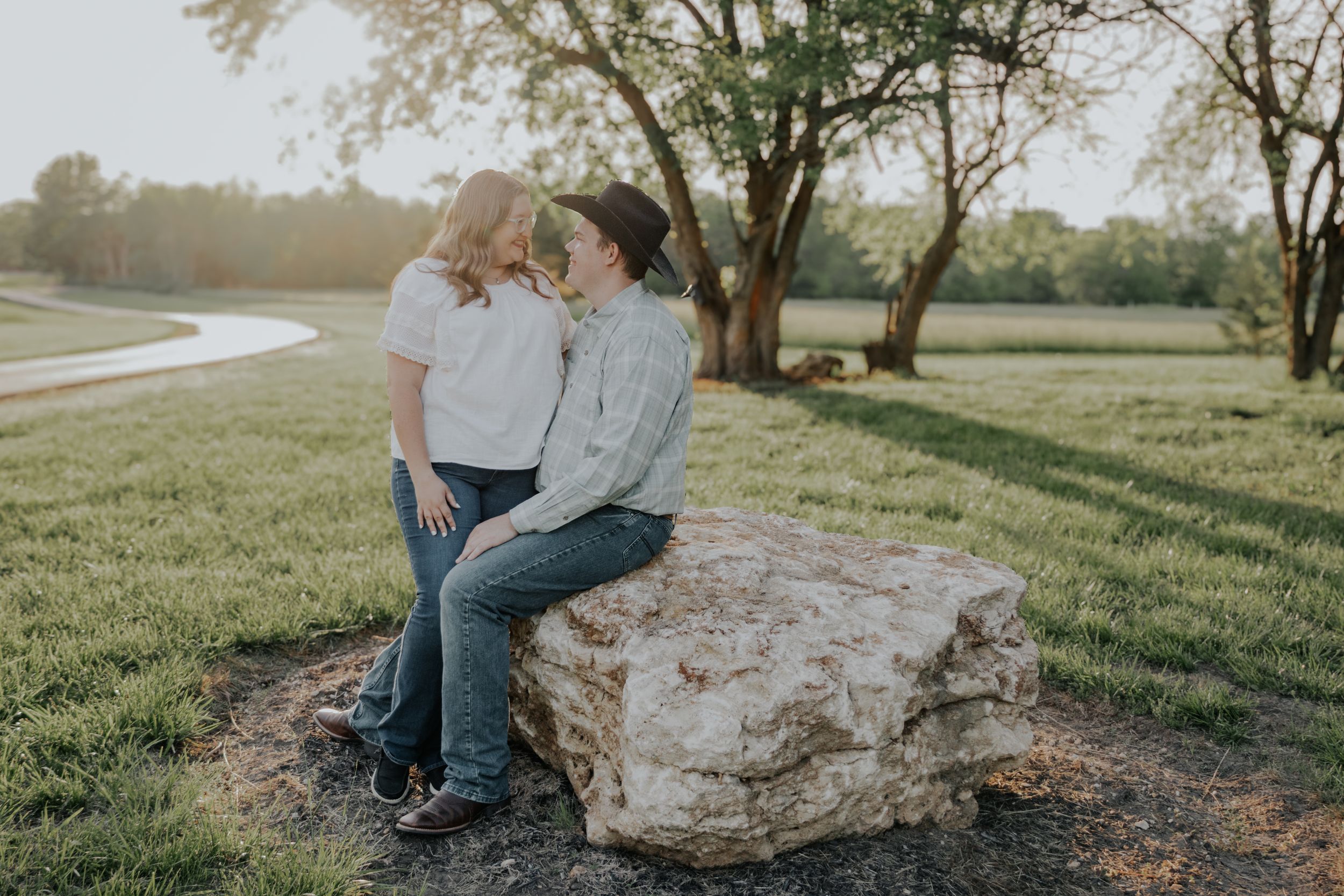 A couple shares a romantic moment while sitting together on a large rock in a sunny park setting.