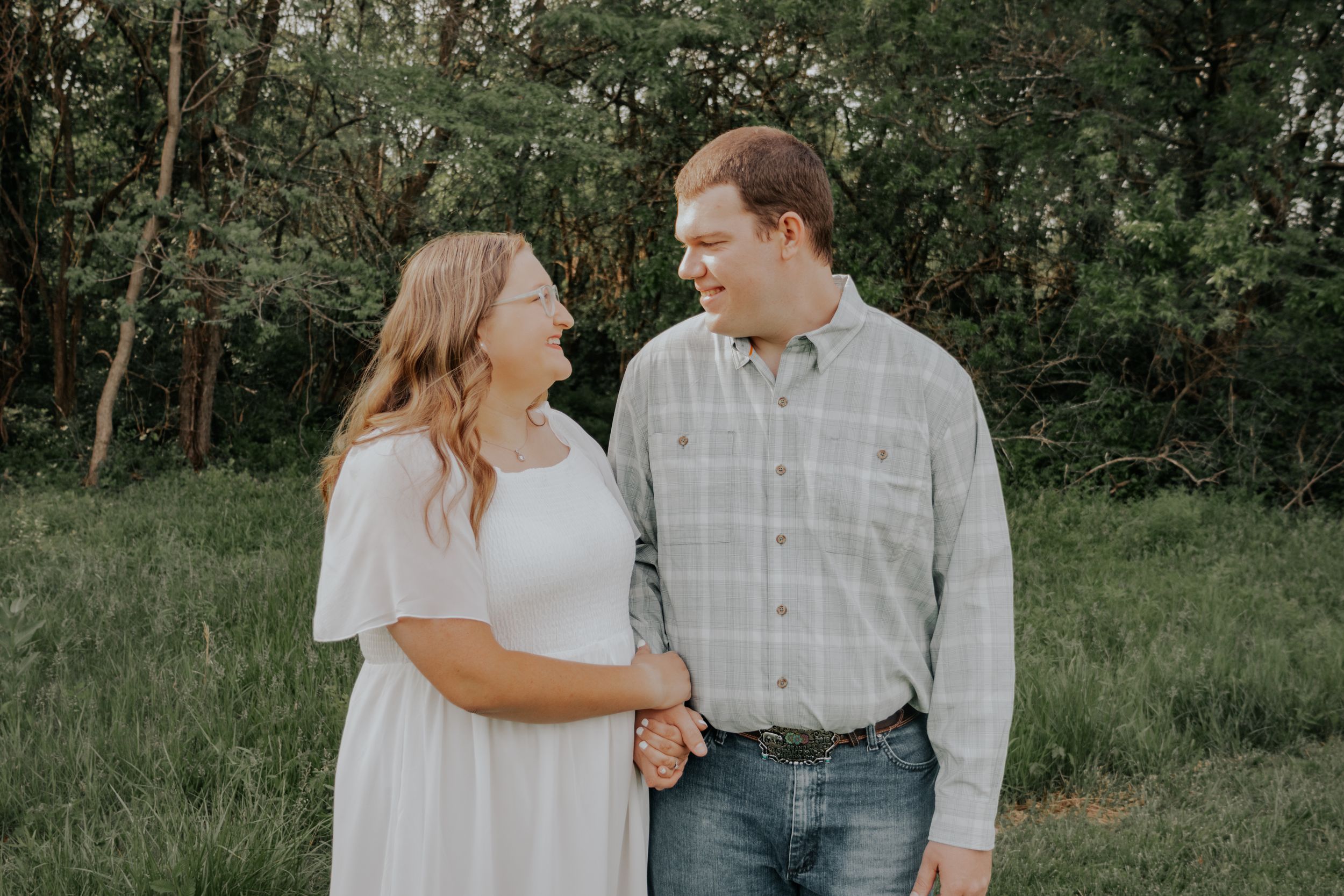A couple in casual attire shares a sweet moment together in a lush green outdoor setting.