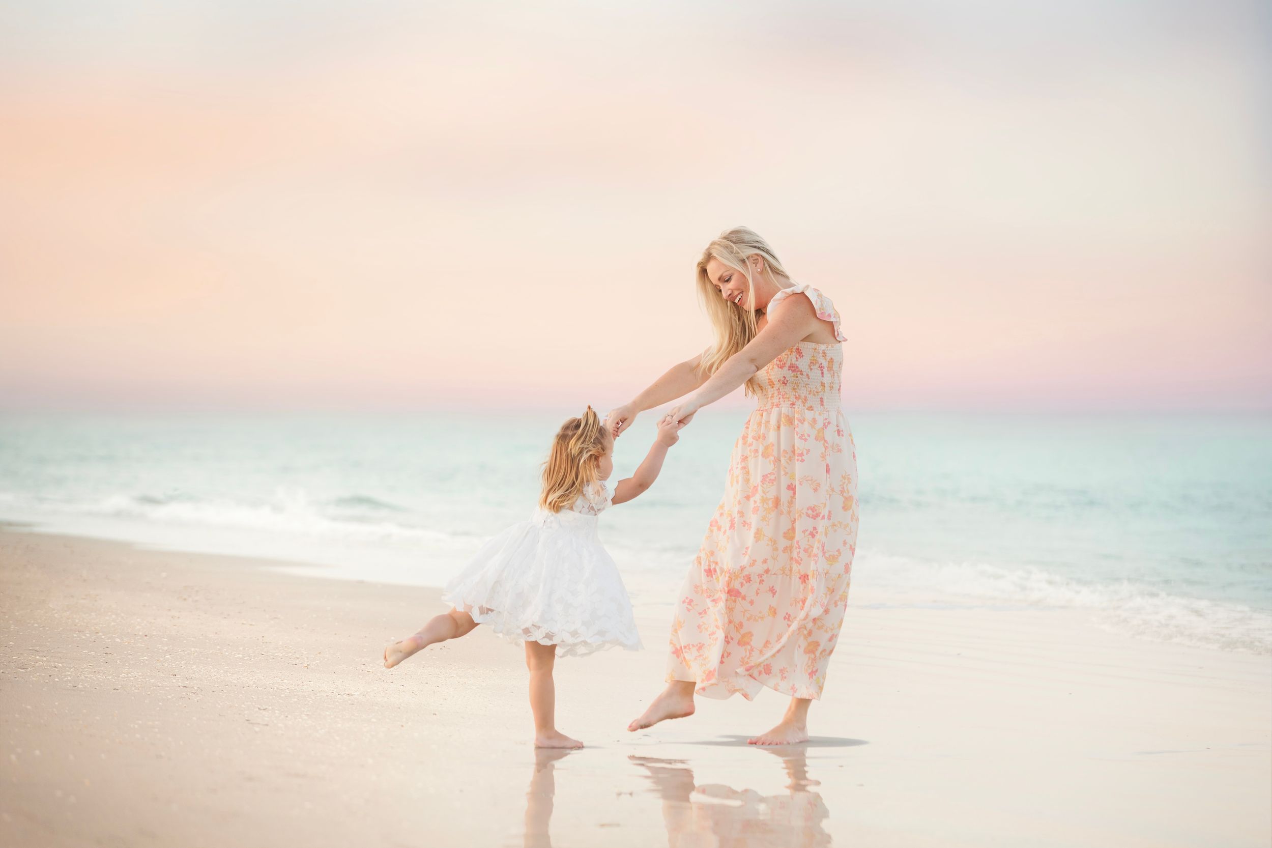 Adult and child playing and twirling together on beach at sunset in soft pink dresses.