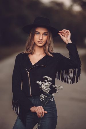 Woman in black fringe jacket and wide brim hat poses with dried flowers.