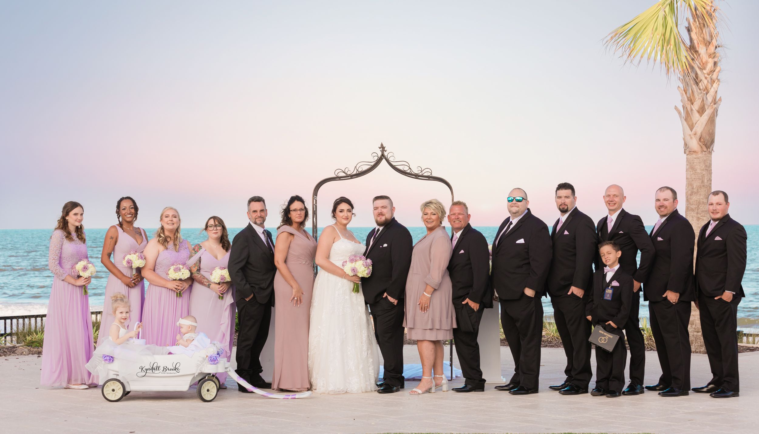 Wedding party in pink and black formal attire poses under palm tree by oceanfront.