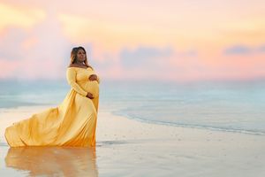 Expectant mother in flowing yellow dress poses on beach during sunset.
