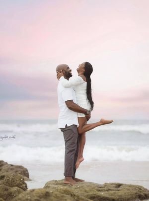 Couple in white clothing shares romantic pose on coastal rocks at sunset.