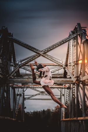 Ballet dancer leaping gracefully in front of an industrial bridge at sunset.