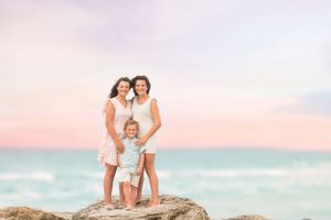 A family stands together on coastal rocks during sunset with pink and turquoise ocean backdrop.