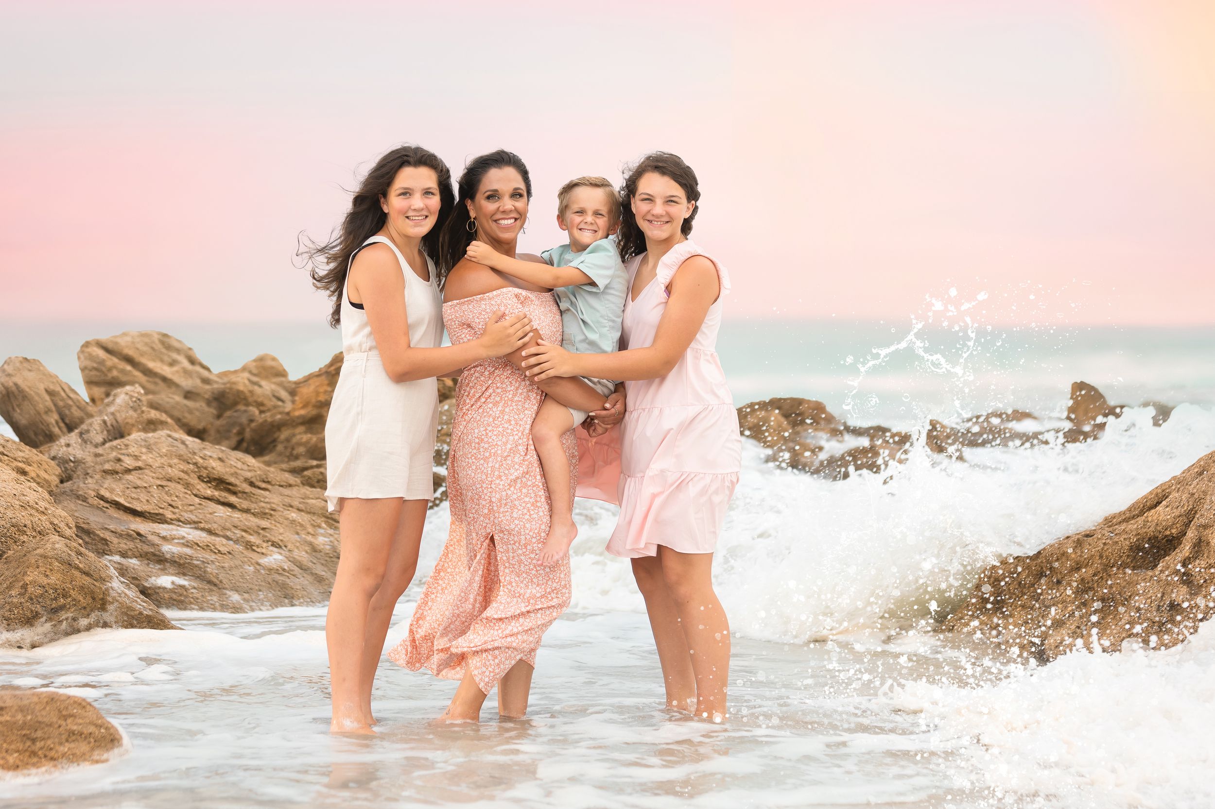 Group of friends in coordinating pink and white outfits pose together on rocky beach.