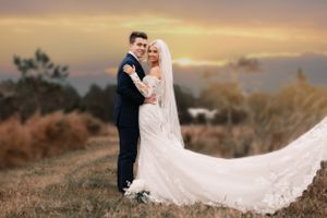 Wedding couple embraces on hilltop with flowing white dress veil at sunset.