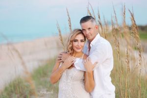 Couple posing in intimate embrace against coastal dunes and sea grass.