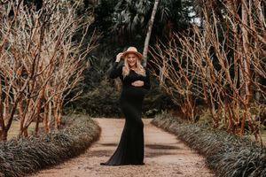 Woman in a black formal dress poses on garden path surrounded by bare branches.