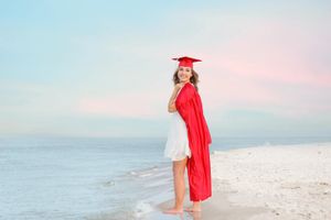 Graduate in red cap and gown poses on sandy beach at sunset.