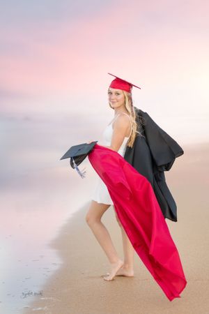 Graduate in white dress poses playfully with graduation gown on beach at sunset.