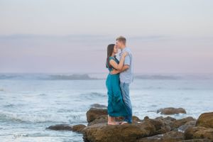 A couple shares an intimate moment while standing on coastal rocks with ocean waves crashing behind them.
