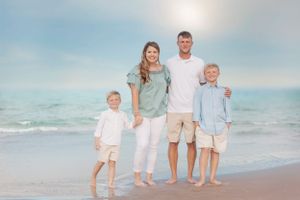 Family of four wearing matching light colors poses together on beach at sunset.