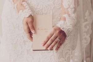 Hands in white lace sleeves hold a blush pink wedding vows book with delicate ring details.