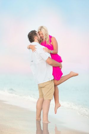 Romantic beachside moment as couple embraces with soft pastel sky background.