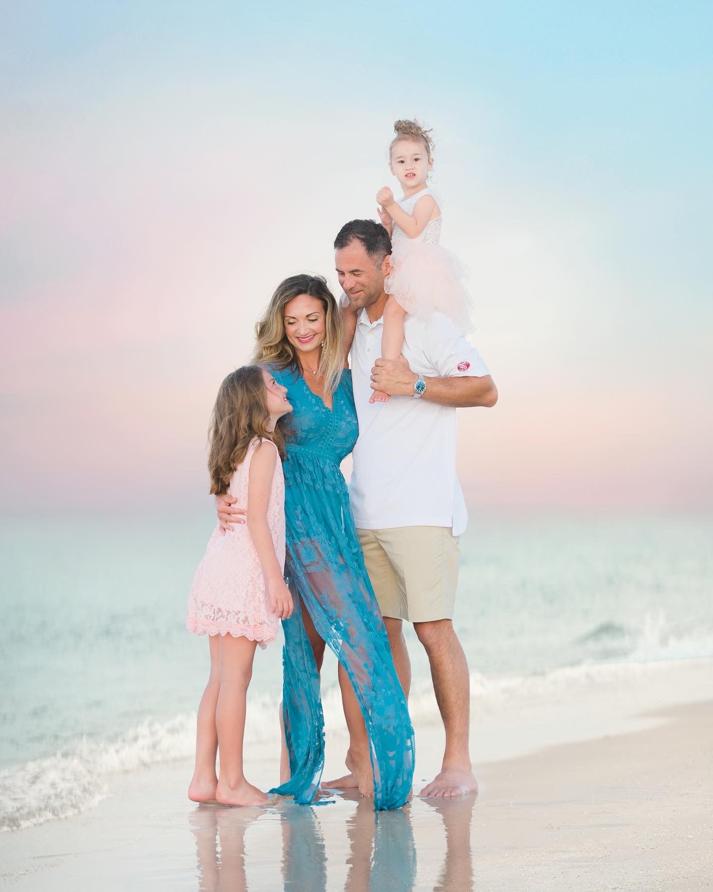 Family of four poses together on beach at sunset with pastel sky background.