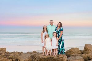 Family in coordinated outfits poses together on coastal rocks during sunset.