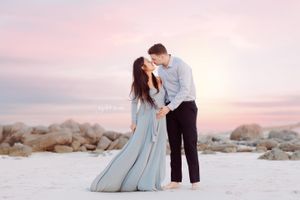 A romantic couple in formal wear embraces on a rocky beach during sunset.