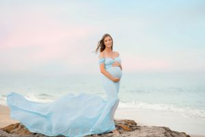 Maternity photo of woman in flowing blue dress on beach rocks during sunset.