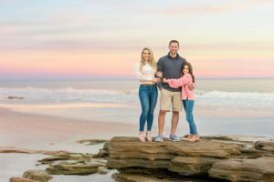 Family standing together on rocky coastal outcrop during sunset beach photo session.