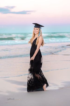 Graduate in black lace dress and cap poses on beach during sunset.