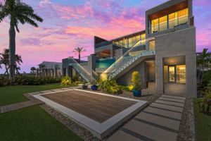 Modern luxury home exterior with illuminated staircase and palm trees under vibrant sunset sky.