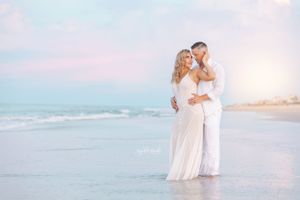 Couple in white outfits sharing intimate moment on dreamy beach at sunset.