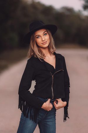 Stylish woman in black fringe jacket and hat poses on outdoor path at dusk.