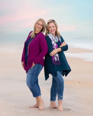 Two friends laughing together on beach at sunset wearing cozy sweaters.