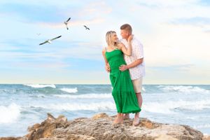 Couple embracing on rocky beach overlook with seagulls flying overhead at sunset.