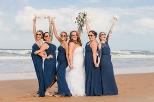 Bridal party in navy blue dresses celebrates joyfully on beach holding bouquets.