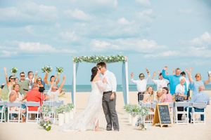 Beach wedding ceremony under white arch with excited guests cheering for the newlyweds.