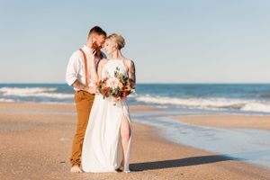 Wedding couple embracing on sandy beach holding coral and peach bouquet with ocean waves in background.