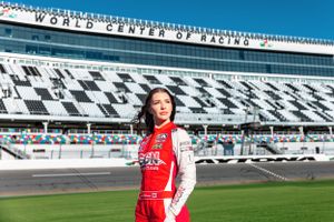 A person in racing suit stands in front of Daytona International Speedway's iconic grandstand.