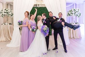 A wedding party poses playfully in front of a floral wall with purple bridesmaid dresses.