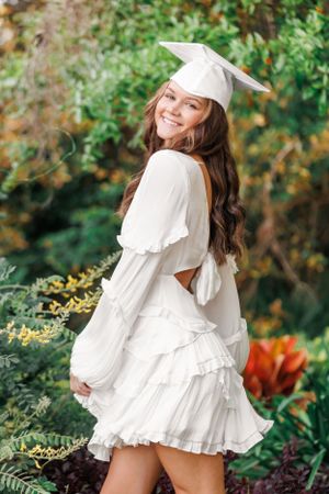 Graduate in white dress and cap twirls in garden setting with autumn foliage.