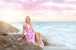 Coastal portrait of someone wearing a pink dress sitting on rocky beach at sunset.