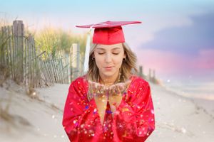 Graduate in red cap and gown blowing glitter from hands on beach at sunset.
